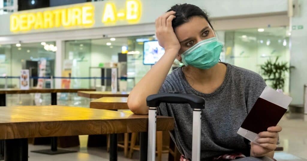 Employee wearing a face mask sitting in an airport departure lounge holding a passport and looking unwell.